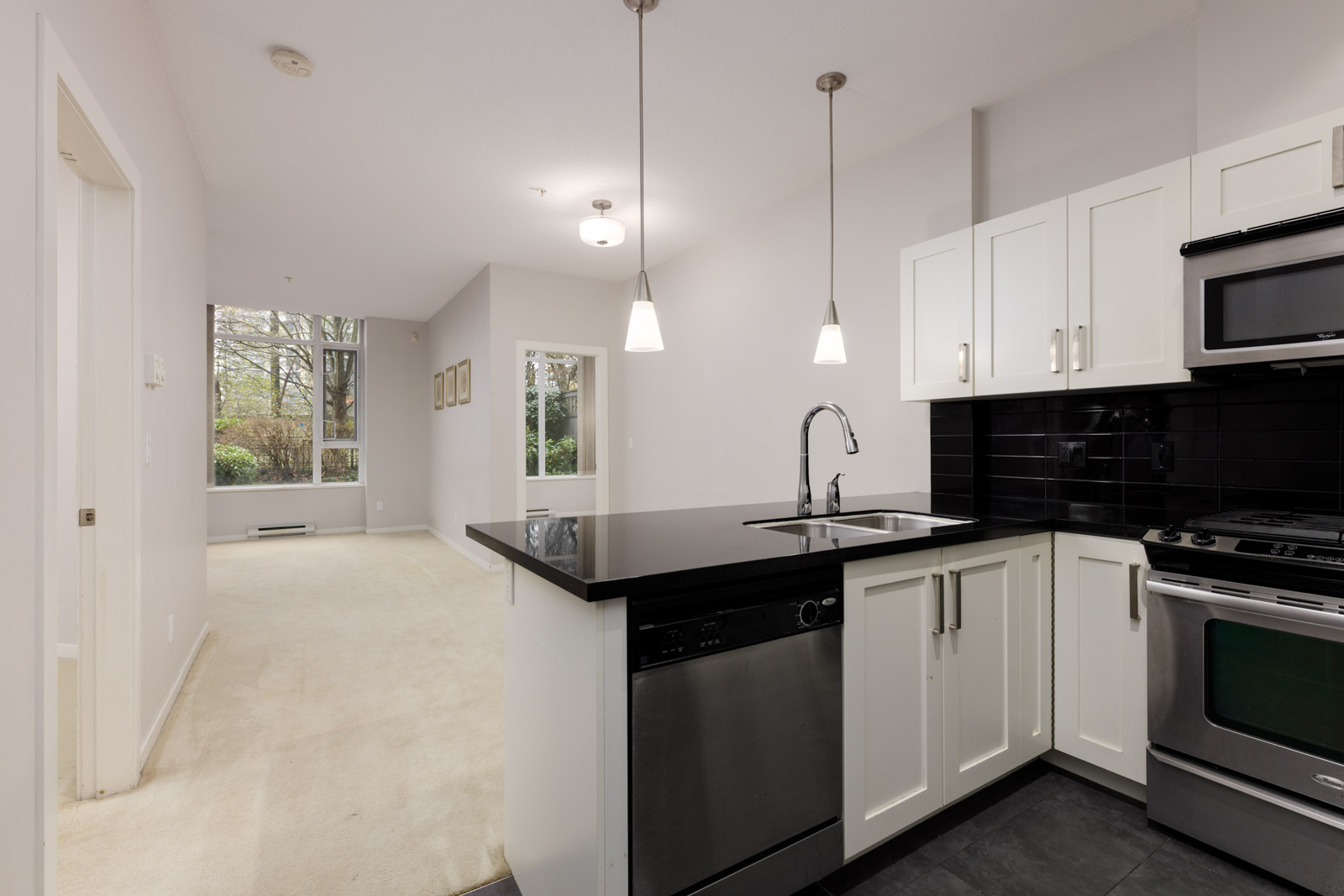 Modern kitchen with black countertops, stainless steel appliances, and white cabinets opens to a carpeted living area with large windows and natural light.