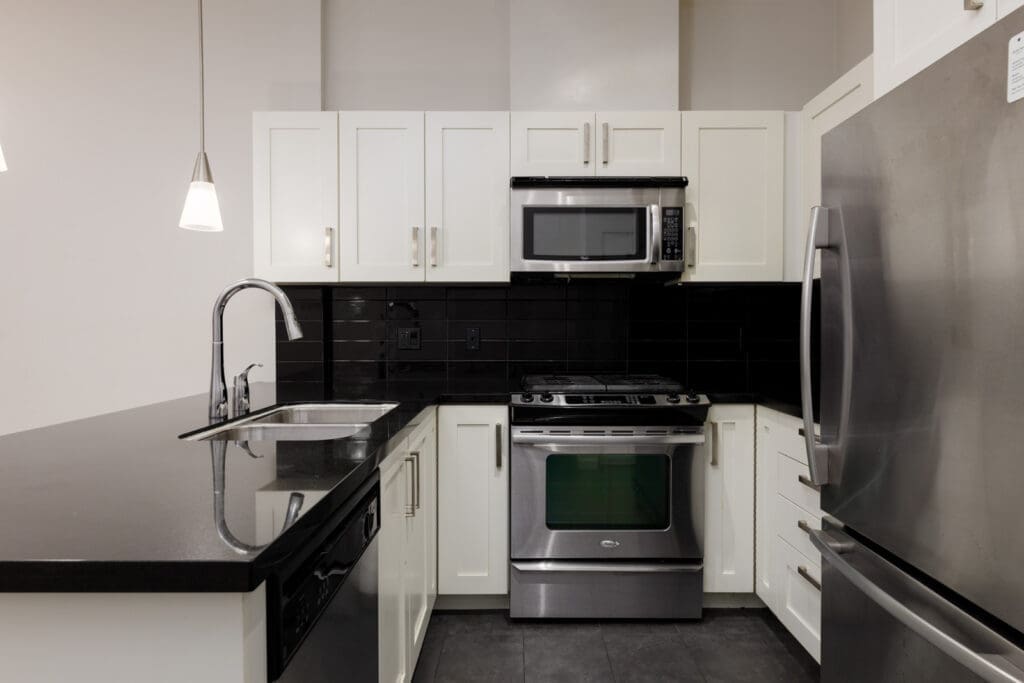 Modern kitchen with white cabinets, black countertops, stainless steel appliances, a built-in oven, microwave, refrigerator, and a double sink under pendant lights.