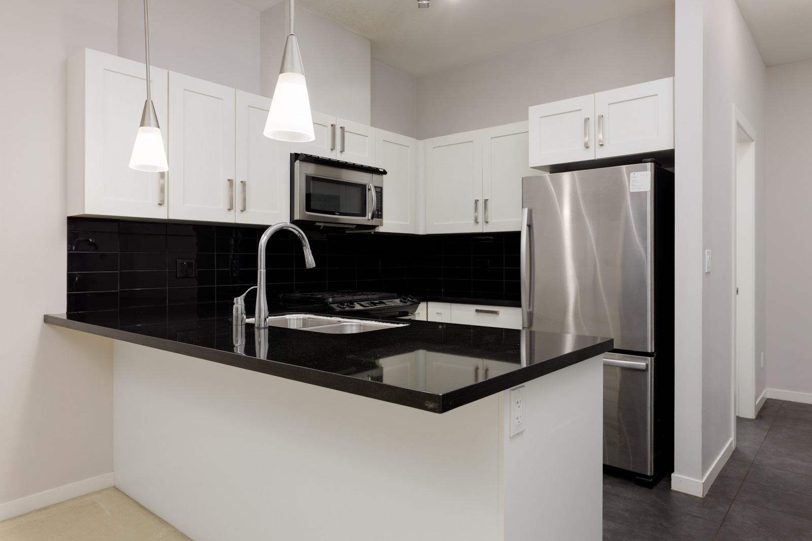 Modern kitchen with white cabinets, black countertops, stainless steel appliances, black tile backsplash, and pendant lighting over a peninsula sink.