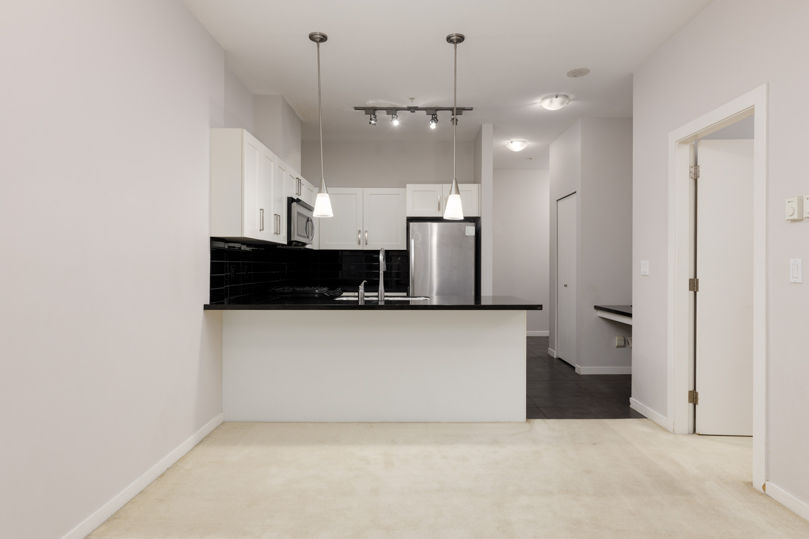 Modern kitchen with white cabinets, black countertops, stainless steel appliances, and pendant lights. Adjacent open space with light carpet flooring.