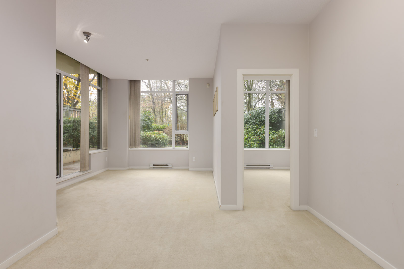 Unfurnished room with light-colored carpet, large windows, and neutral walls; outside greenery visible through the windows.