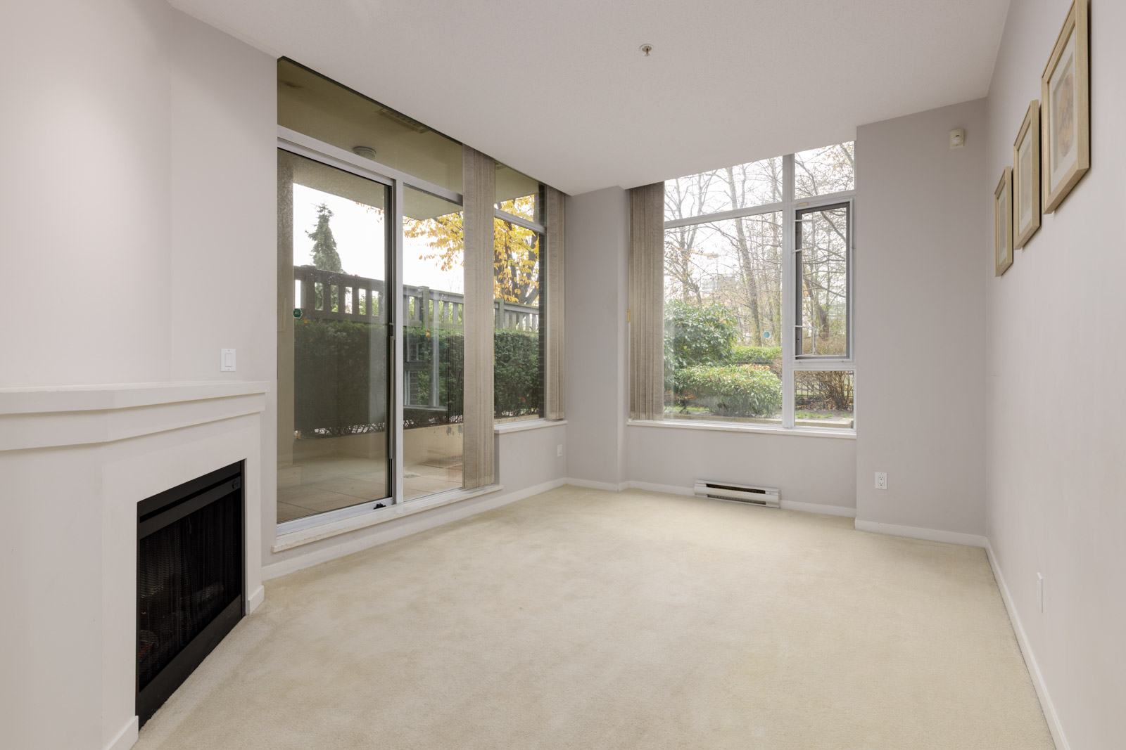 Unfurnished living room with beige carpet, a fireplace, large window, glass door to patio, and neutral-colored walls with framed pictures.