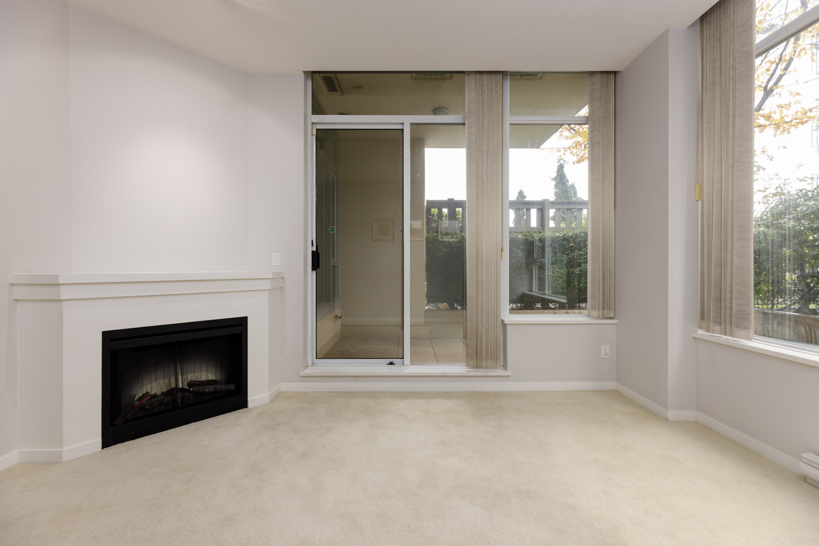 Unfurnished living room with a gas fireplace, beige carpet, large windows, and a sliding glass door leading to a covered patio or balcony.