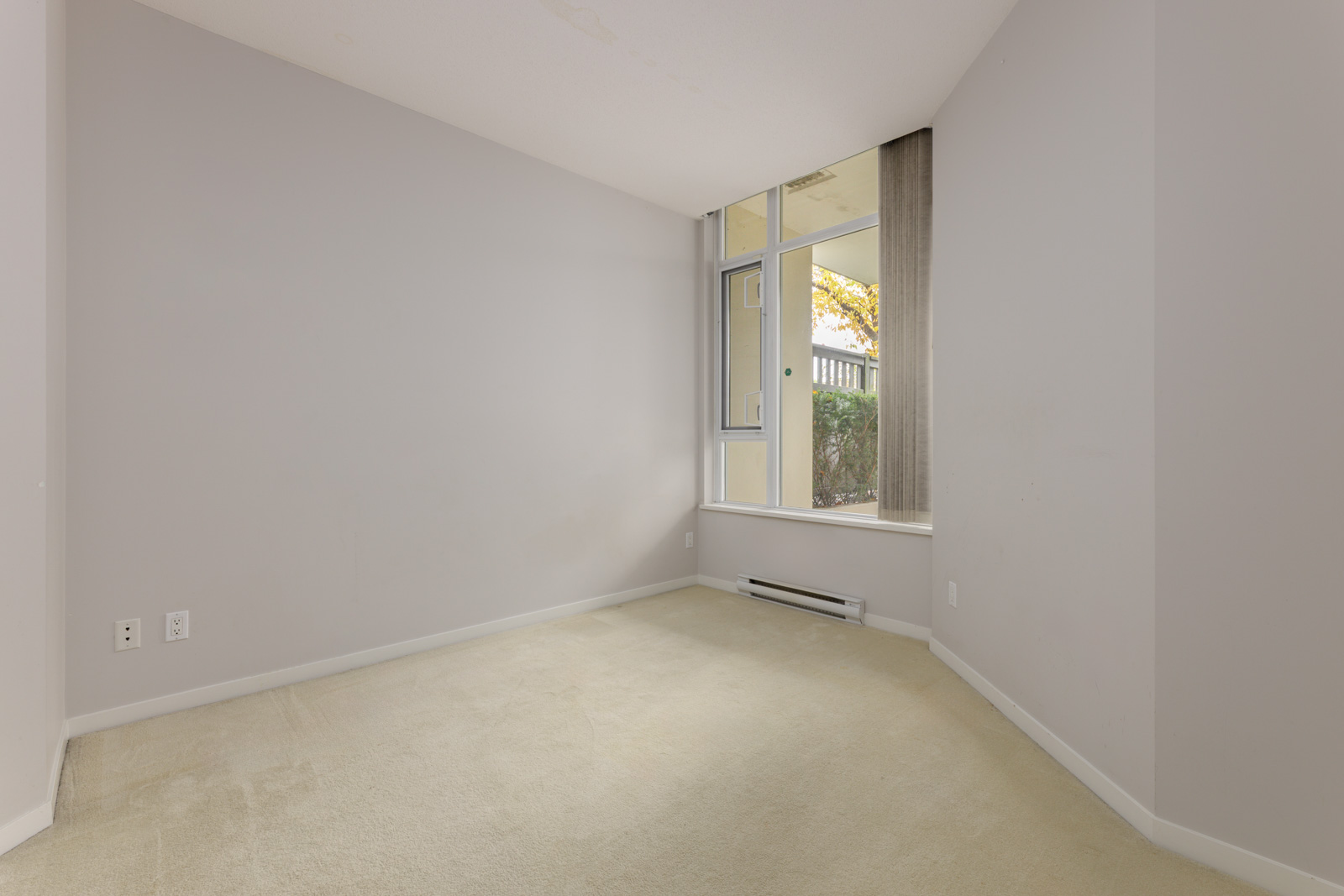 Empty room with beige carpet, light gray walls, and a large window showing an outdoor area with trees and a fence. Electrical outlets are visible on the left wall.