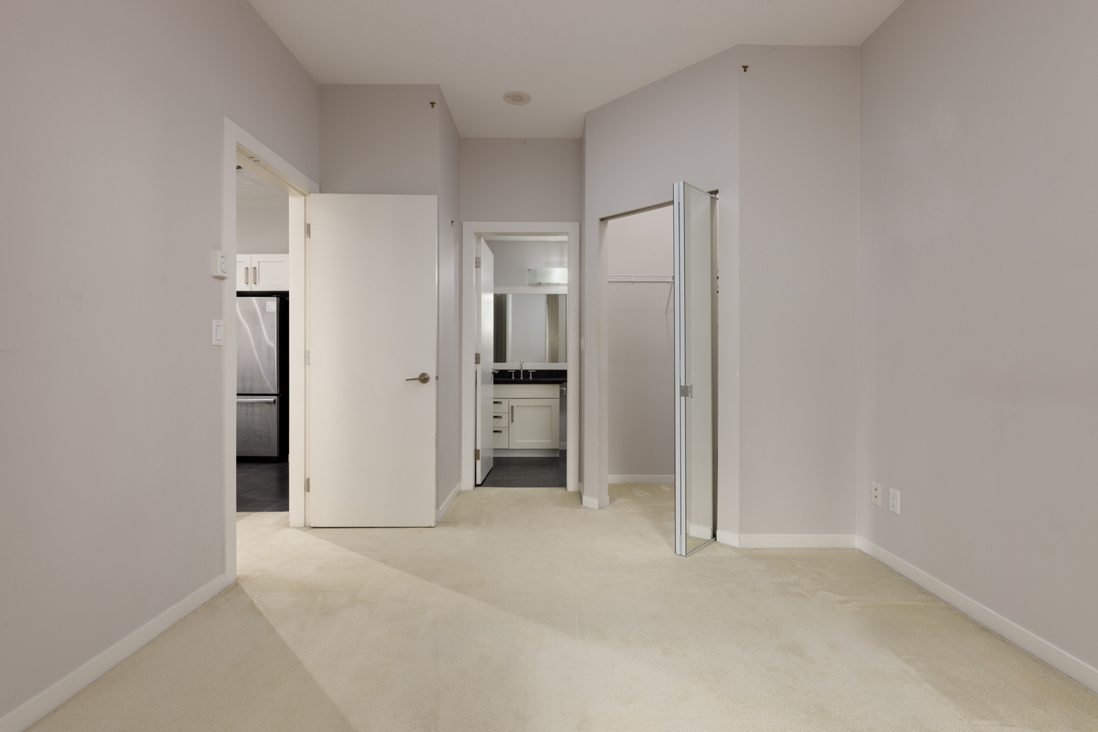 Empty bedroom with light-colored carpet, open door to bathroom, closet with mirrored sliding doors, and light gray walls.
