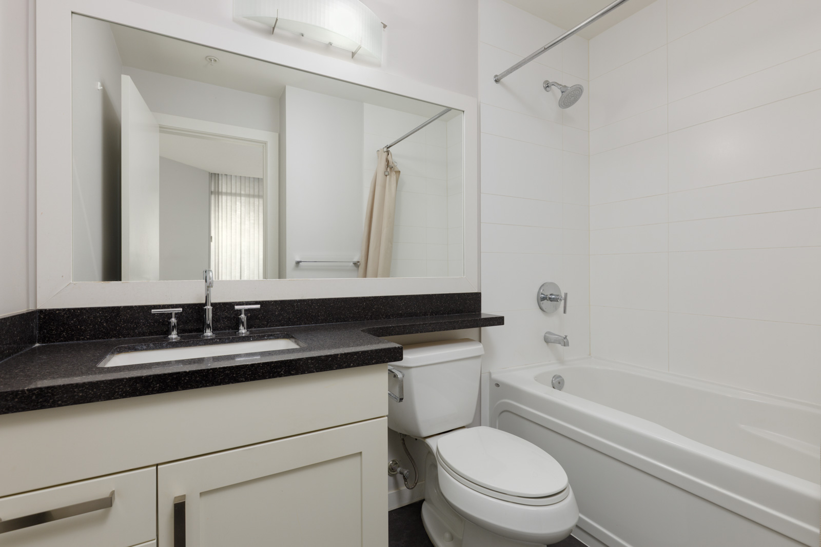Modern bathroom with a white bathtub, shower, toilet, sink, large mirror, and black countertop. Light fixture above the mirror; door partially open.