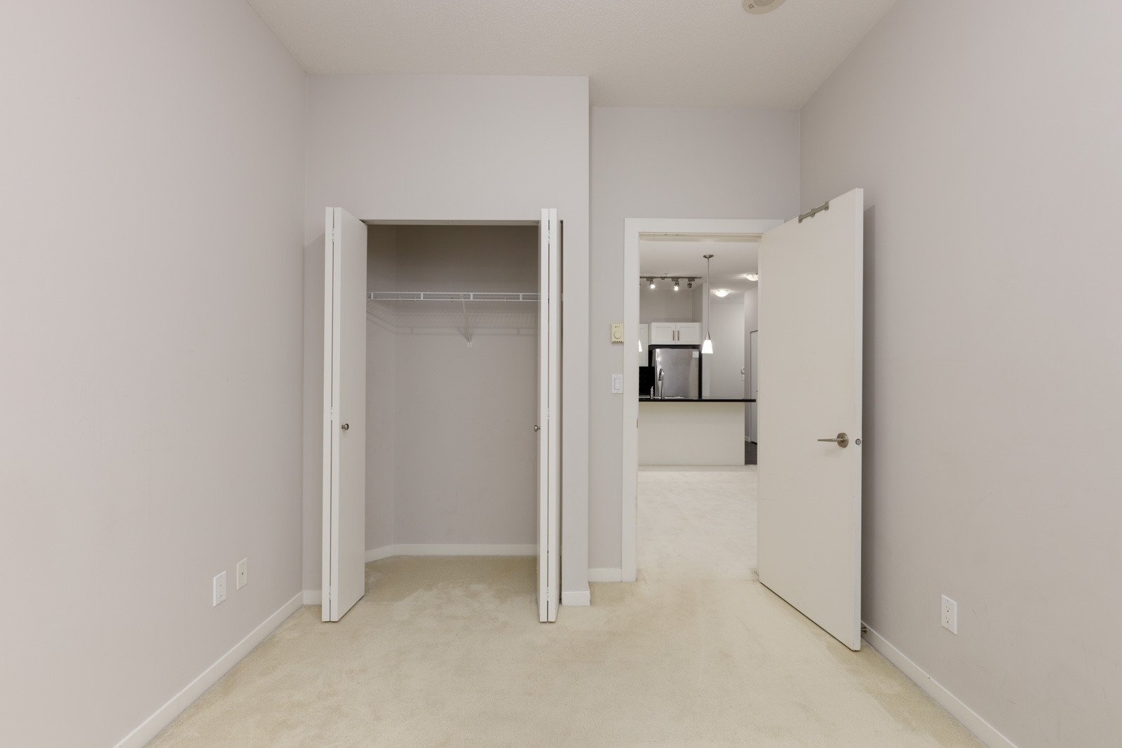 Empty room with beige carpet, open closet doors showing a shelf and hanging rod, and an open door leading to another room with a kitchen in the background.
