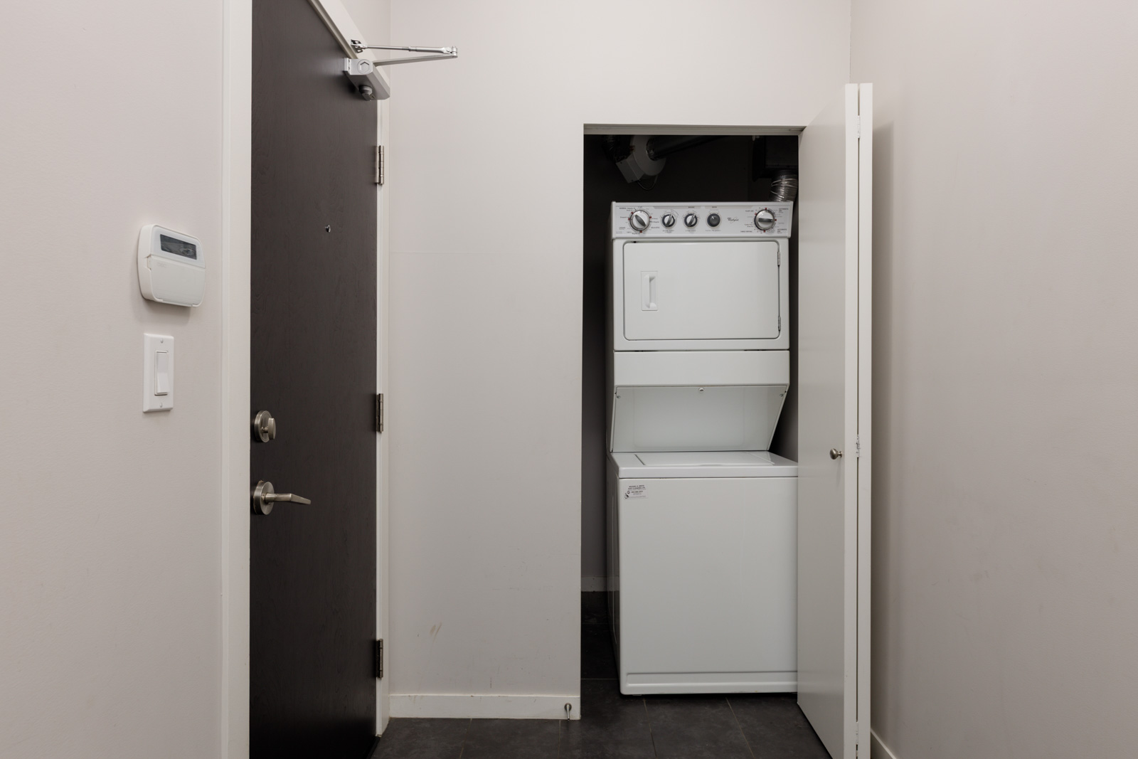 A small laundry area with a stacked washer and dryer is shown in a closet with the door open, next to a black entrance door and a wall thermostat.
