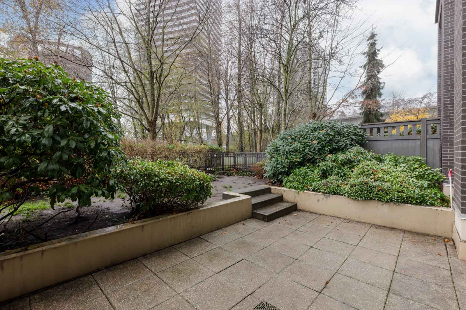 A paved patio with raised planters and green shrubs, bordered by a fence, with tall buildings and bare trees in the background.