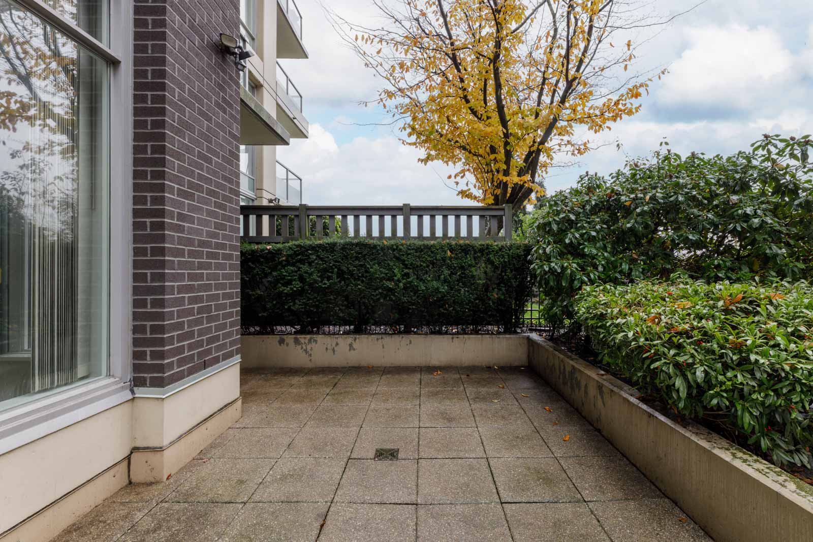 A small outdoor patio with concrete tiles, surrounded by green bushes and a wooden fence, next to a brick building. A tree with yellow leaves is visible beyond the fence.