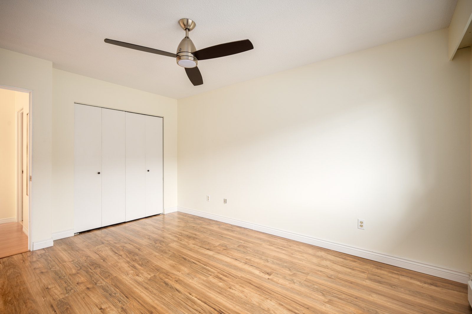 Empty room with light-colored walls, wood flooring, ceiling fan, closed double closet doors, and an open doorway leading to another area.