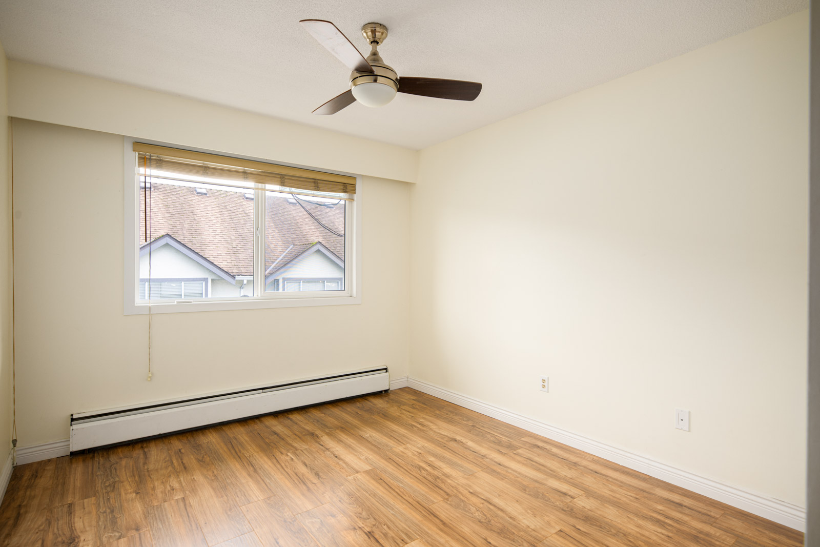 Empty room with light-colored walls, wood flooring, a ceiling fan, a large window with blinds, and a baseboard heater below the window.