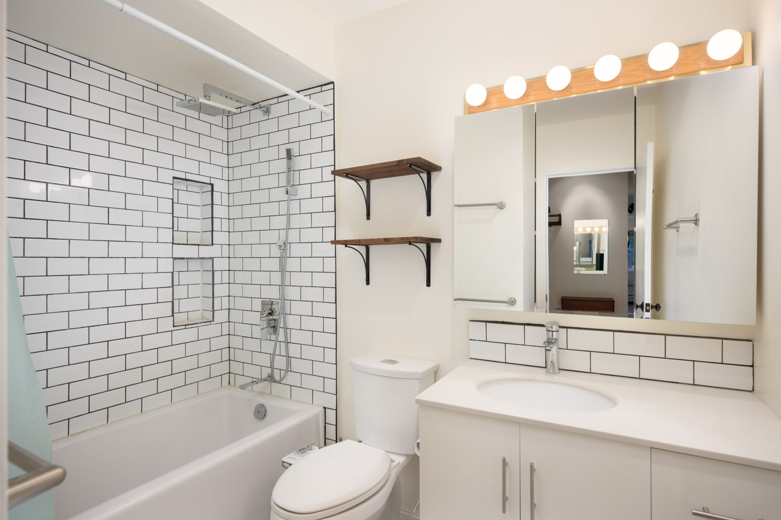 Modern bathroom with white subway tile, bathtub, wall-mounted shelves, toilet, vanity with sink, large mirror, and bright bulb lighting above the mirror.