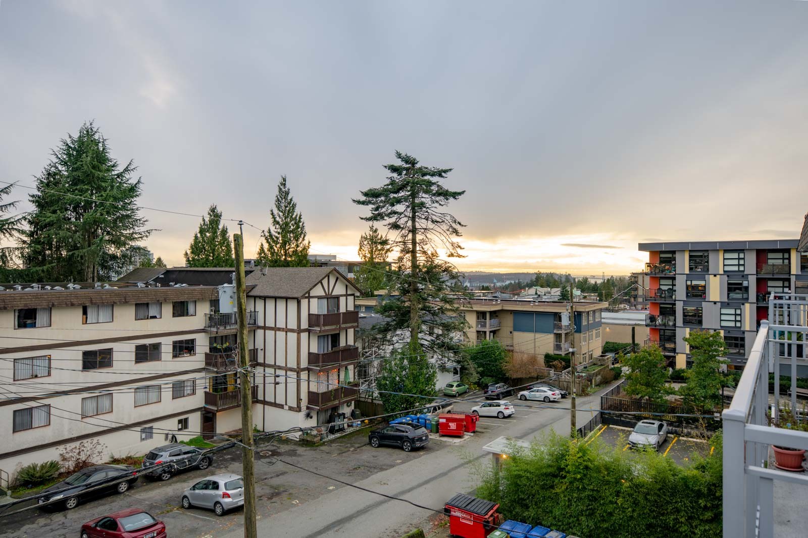 View of a residential area with apartment buildings, parked cars, trees, and a cloudy sky at sunset.