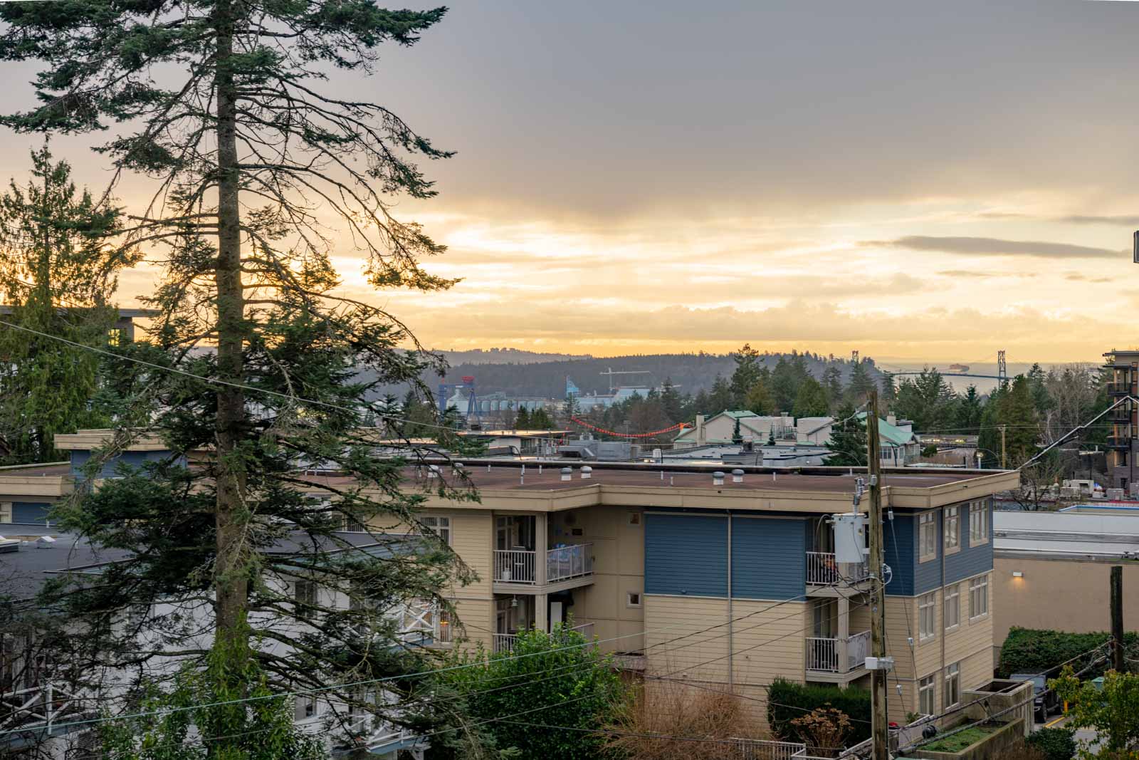 Apartment buildings and trees in the foreground with a view of an industrial area and hills under a cloudy sunset sky in the background.