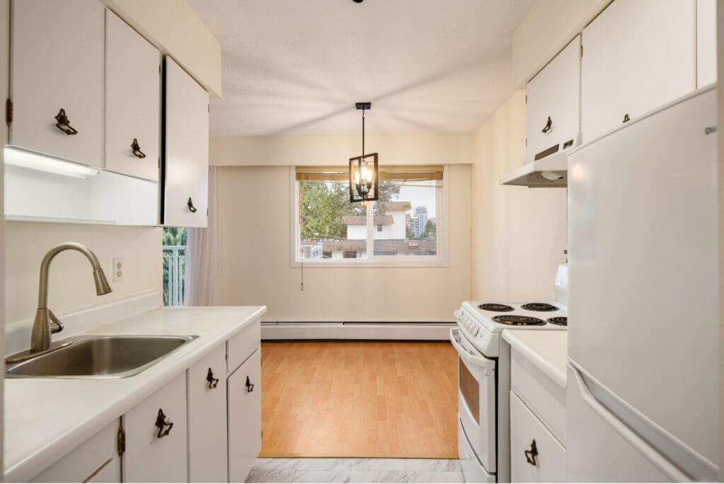 A small kitchen with white cabinets, electric stove, sink, and refrigerator opens into a dining area with wood flooring and a large window.