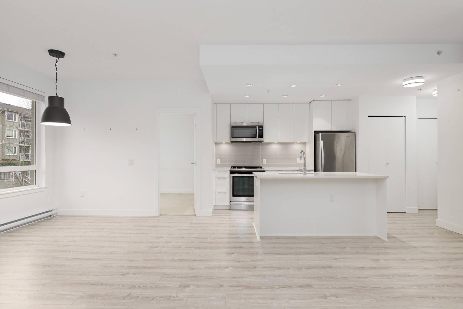 Modern, unfurnished apartment kitchen with white cabinets, stainless steel appliances, island, light wood floors, and large window.