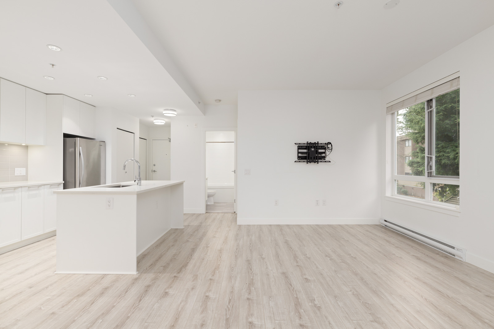 Empty modern apartment with light wood flooring, white walls, open kitchen with island, large window, and wall mount for TV.