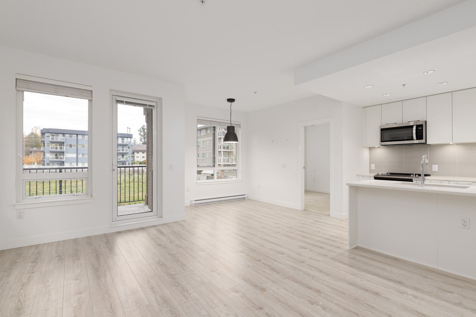 Bright modern apartment with light wood flooring, white walls, large windows, a sliding door to a balcony, built-in kitchen appliances, and a black pendant light fixture.