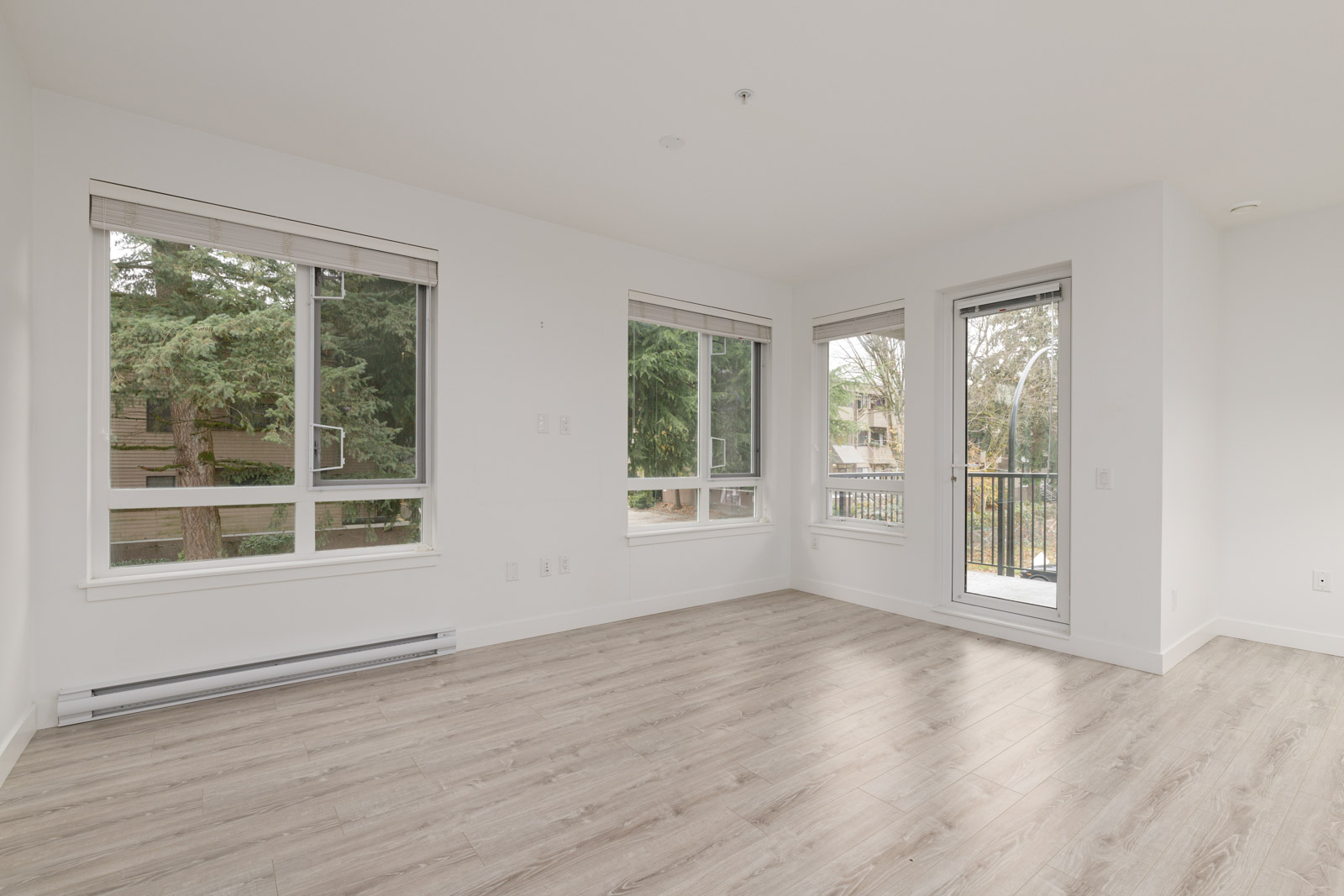 Empty room with light wood flooring, white walls, multiple large windows, and a glass door leading to a balcony; trees visible outside.