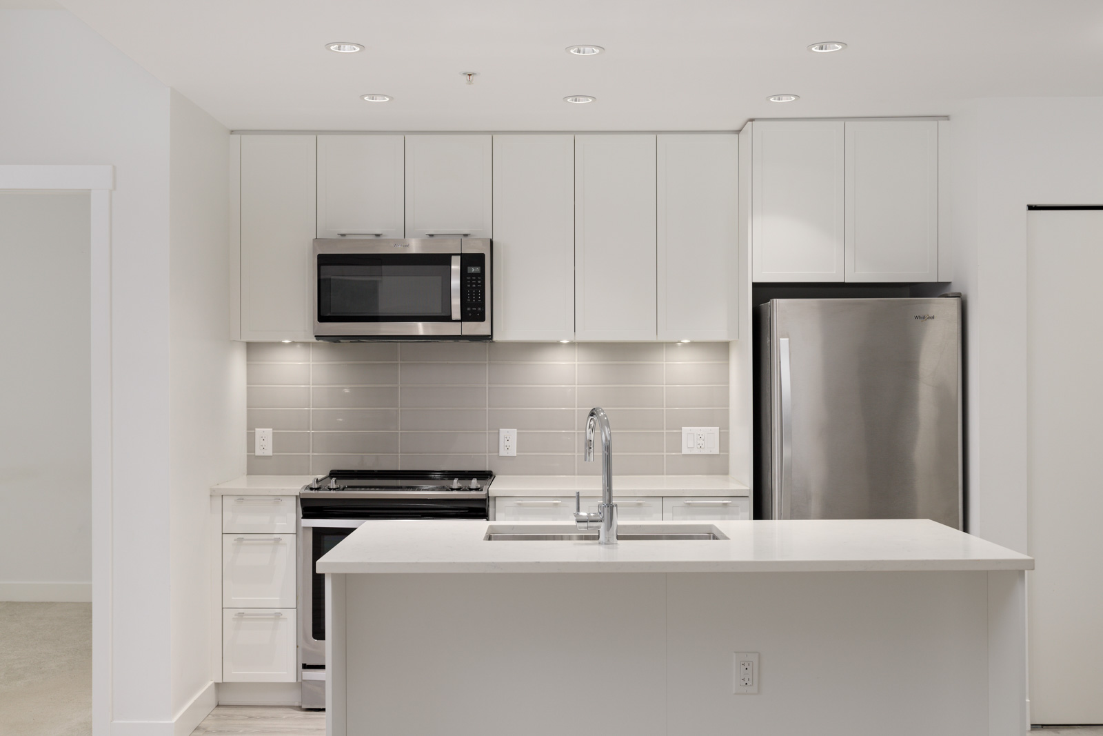 Modern kitchen with white cabinets, stainless steel appliances, gray tile backsplash, built-in microwave, and a sink on a central island.