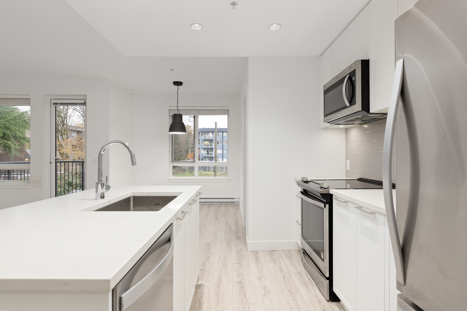 Modern kitchen with white cabinets, stainless steel appliances, an island with sink, large windows, and light wood flooring.
