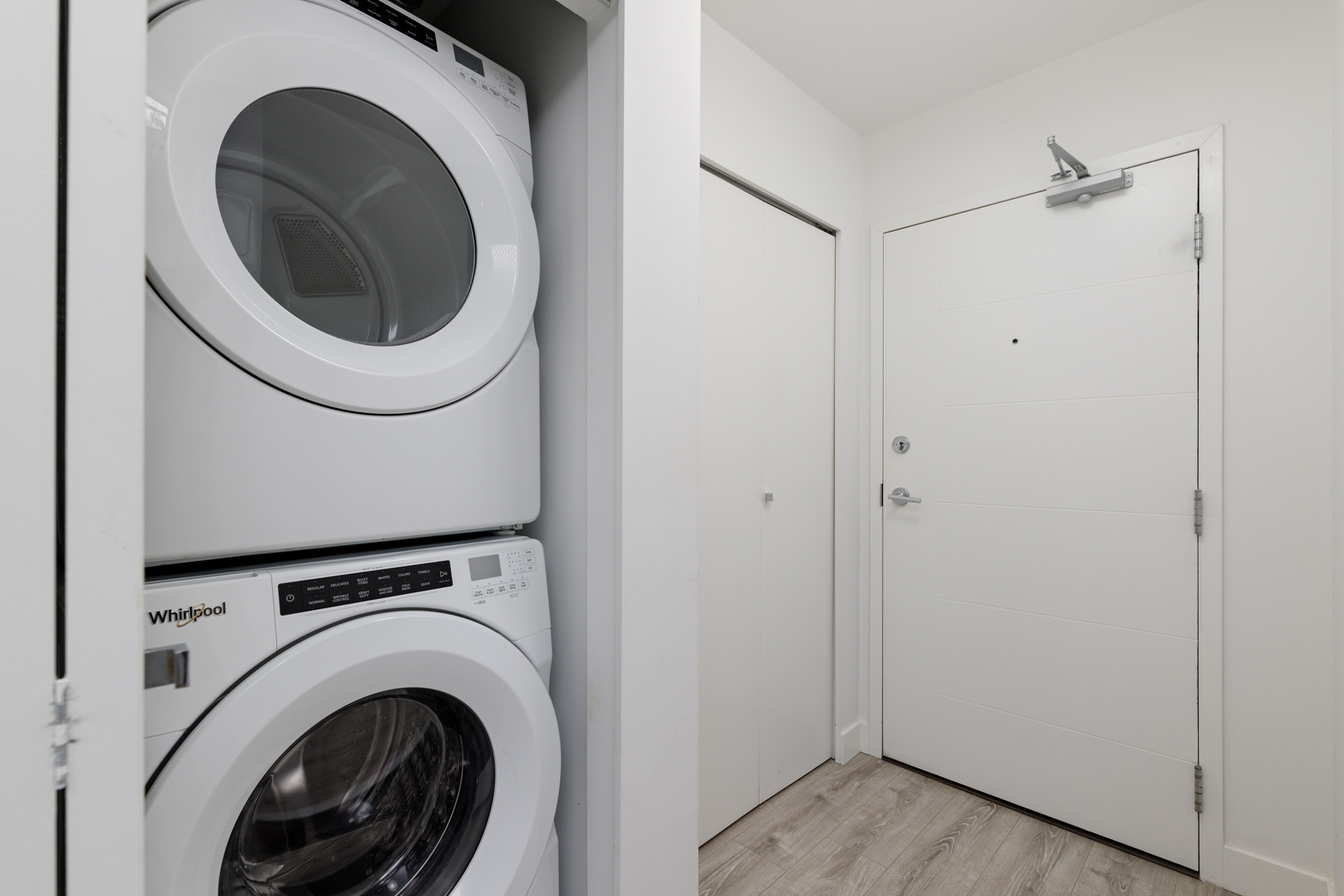 Front-loading washer and dryer stacked in a closet next to a white entrance door in a modern, minimalist laundry area.