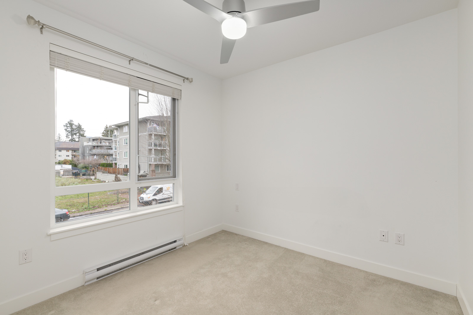 Empty white room with beige carpet, ceiling fan with light, window with blinds, and view of apartment buildings outside.