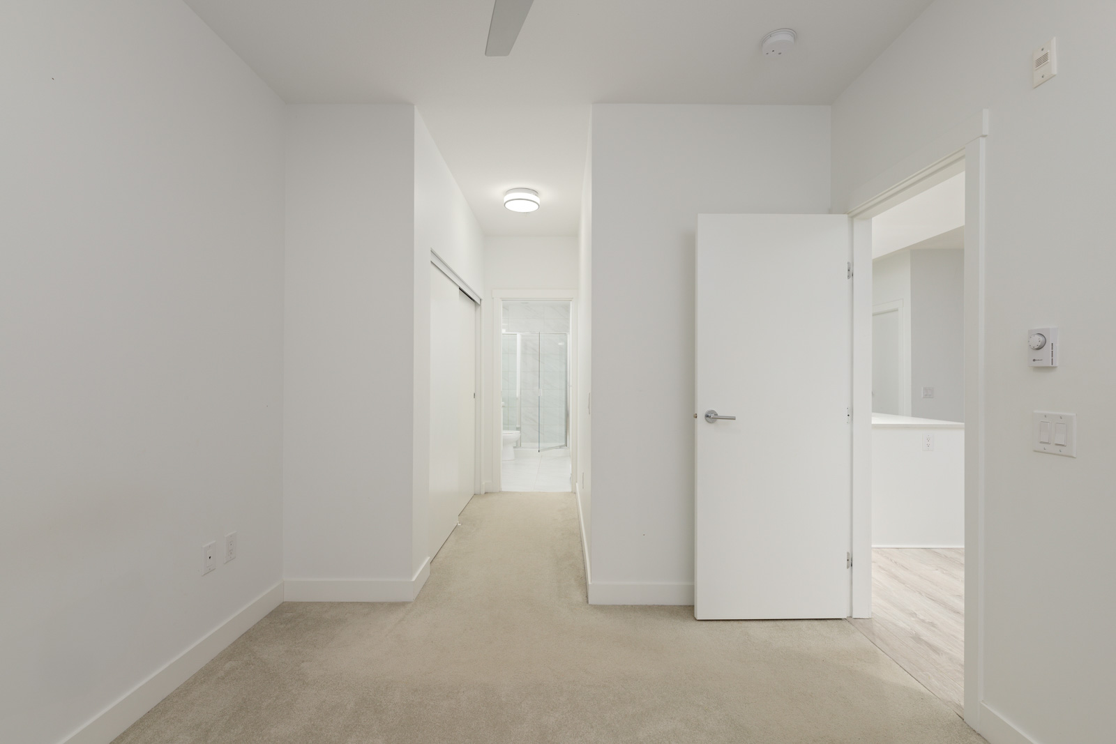 A hallway with beige carpet leads to a bathroom with a glass shower; white walls and doors, and a ceiling light are visible.