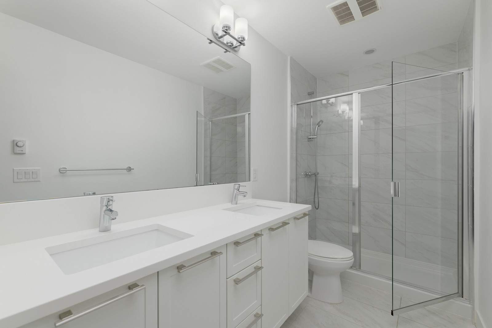 Modern bathroom with double sinks, white cabinets, a large mirror, glass shower enclosure, toilet, and white tile flooring and walls.