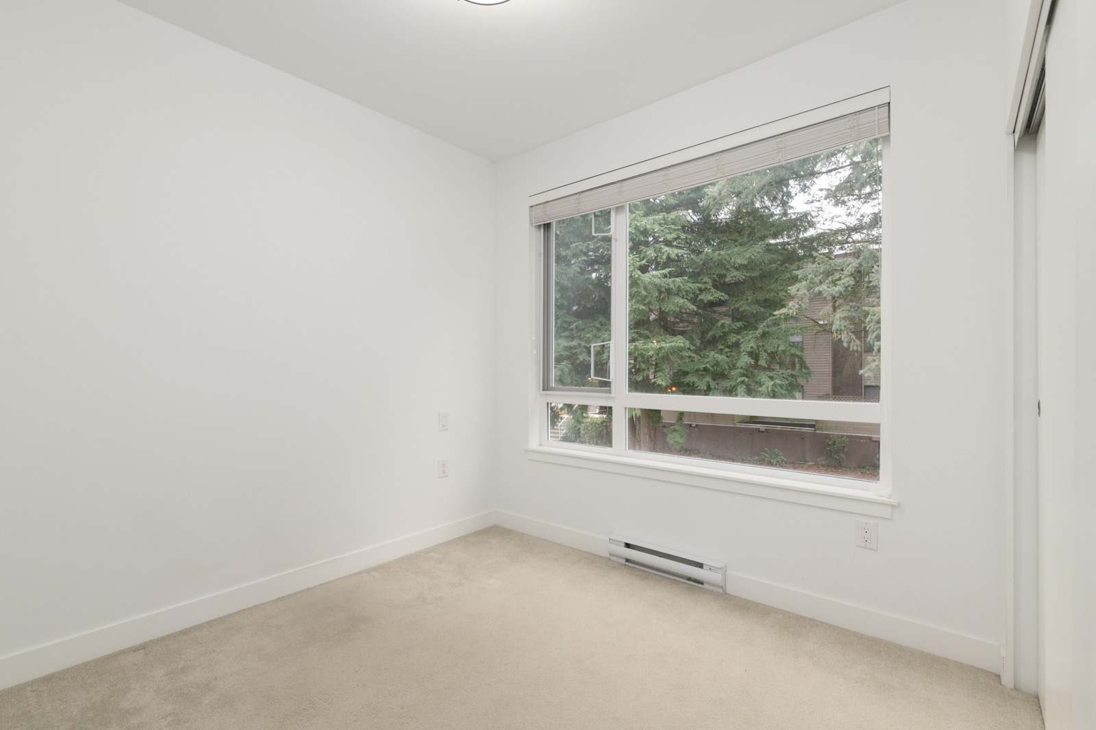 Empty room with white walls, beige carpet, a large window with blinds, and a view of green trees outside.