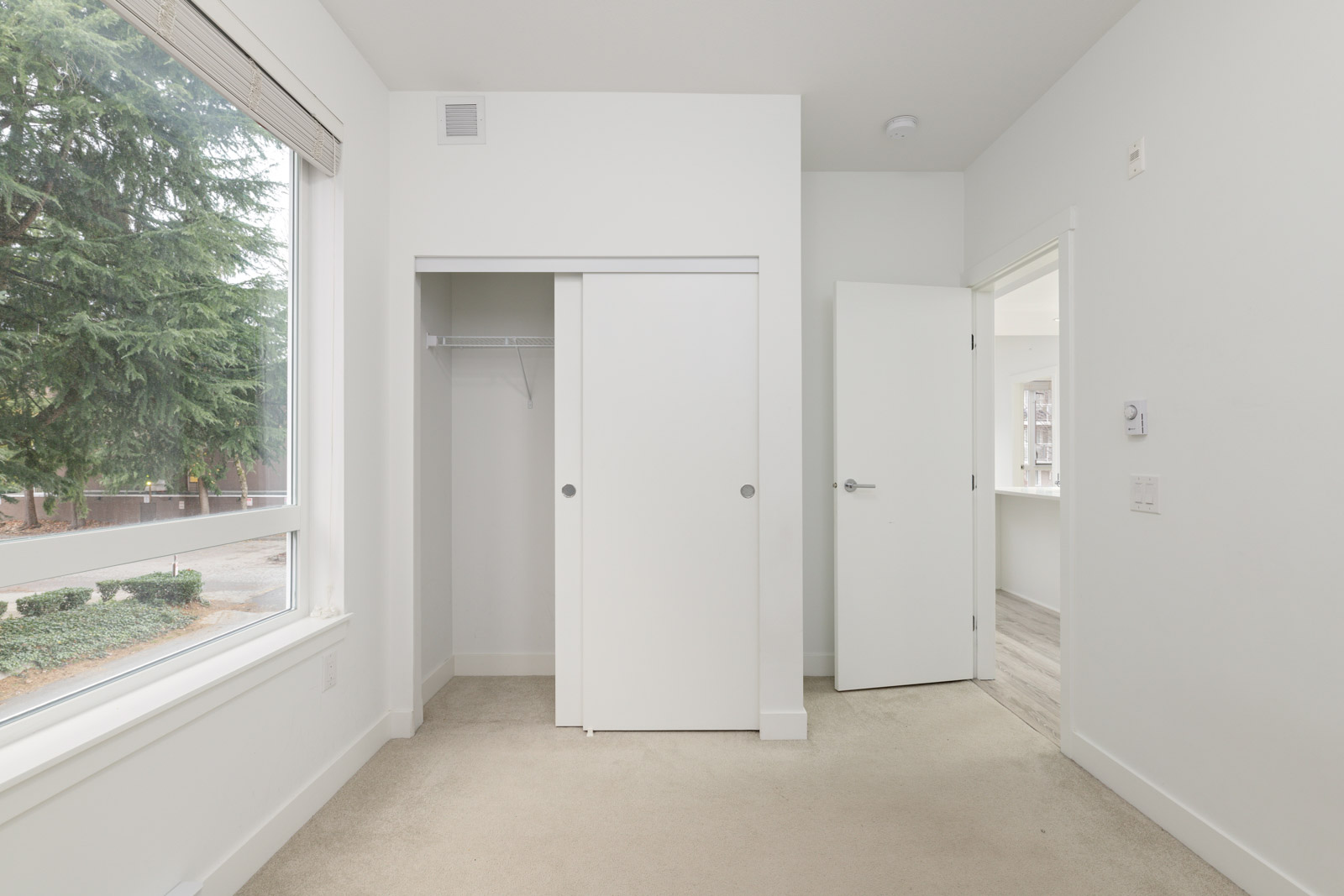 Empty room with beige carpet, white walls, a large window, an open closet with sliding doors, and an open door leading to another area.