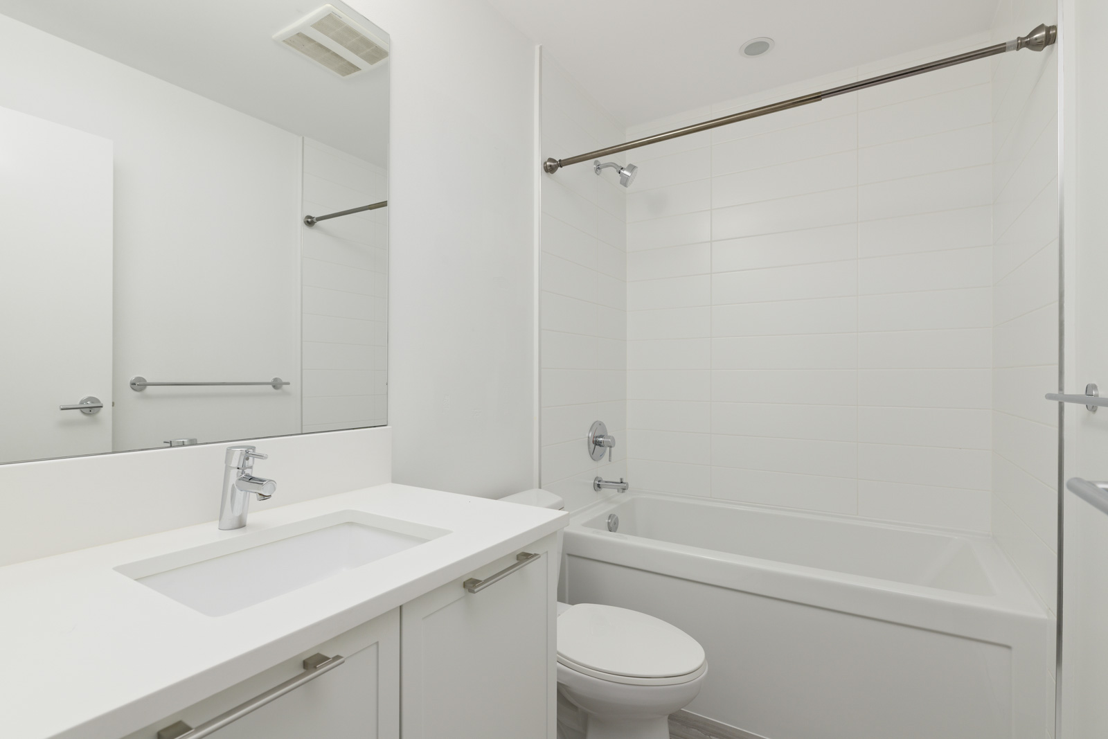 A modern bathroom with white walls, a built-in bathtub with a shower rod, a toilet, and a white vanity with a sink and faucet.