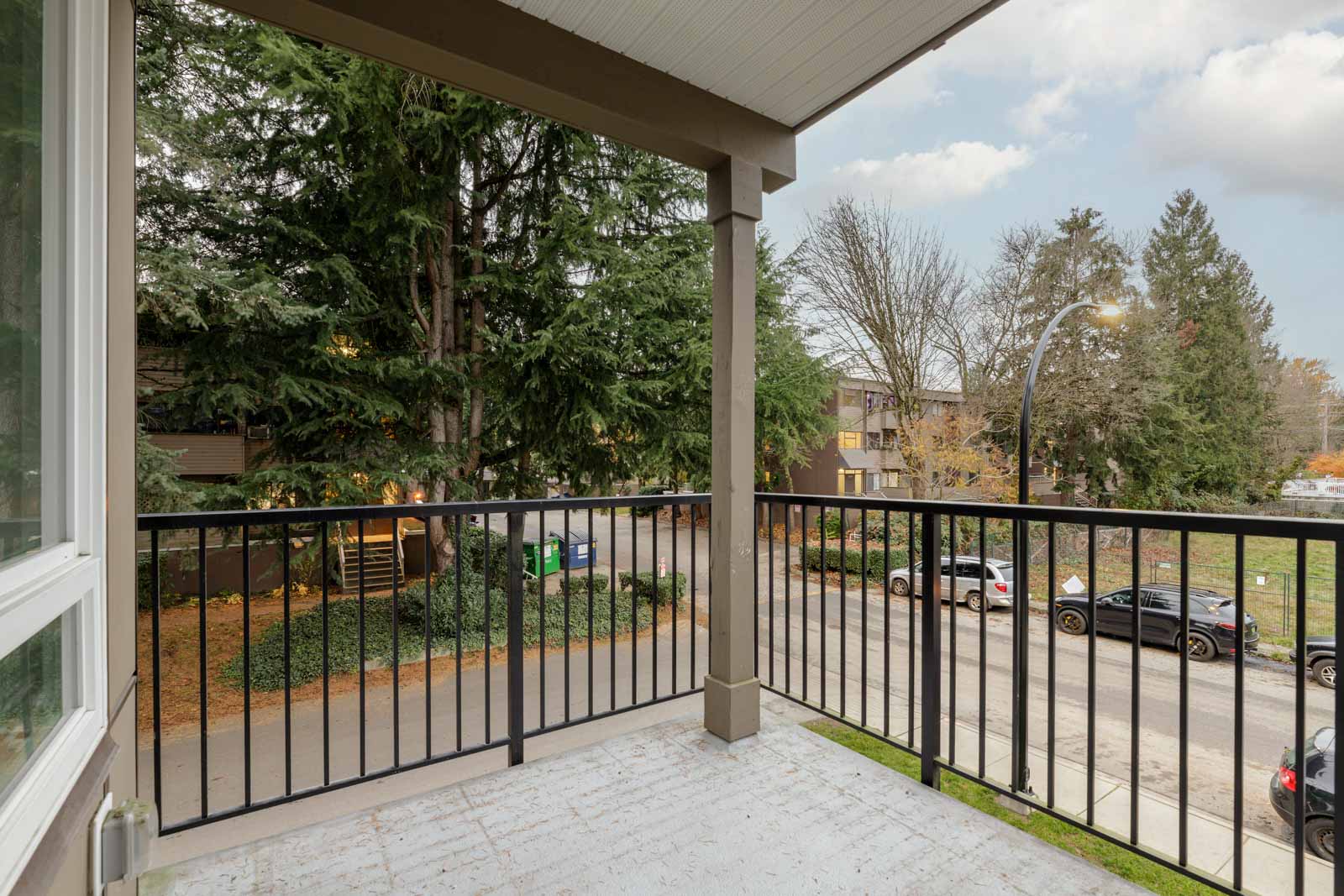 A covered balcony with a black metal railing overlooks a street with parked cars, trees, and nearby apartment buildings on an overcast day.