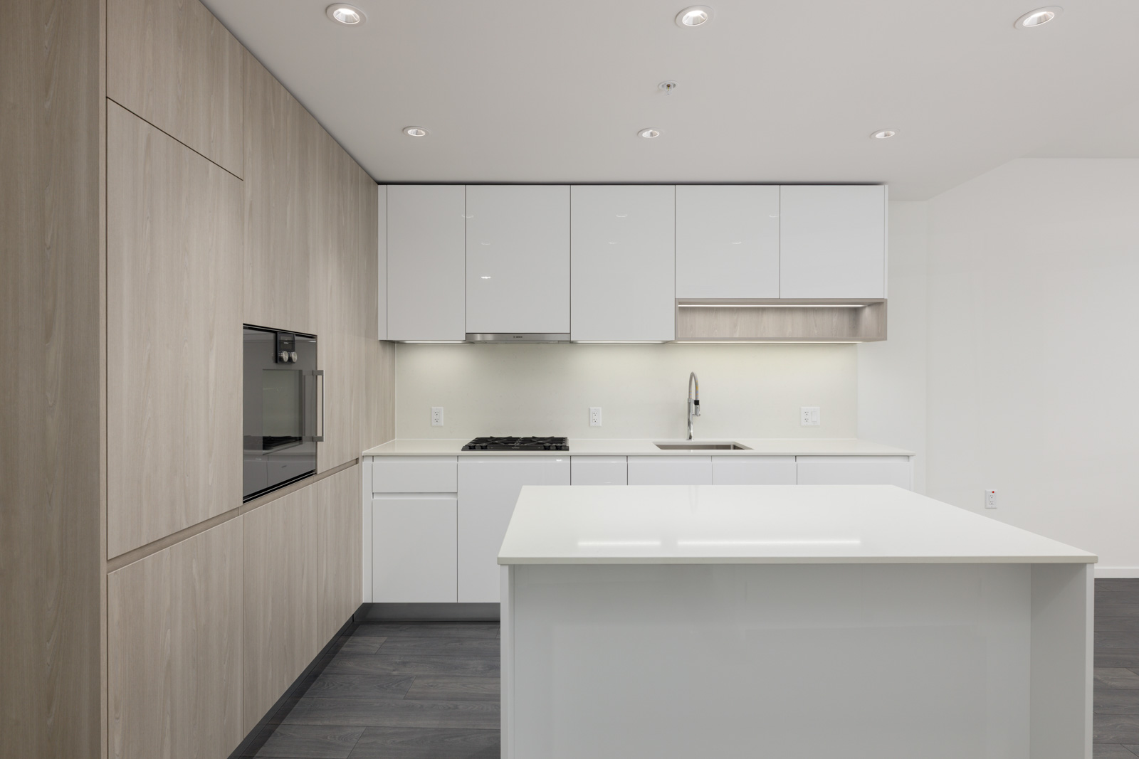 Modern, minimalist kitchen with white cabinets, built-in oven, gas stove, island, and light wood accents; under-cabinet lighting illuminates the workspace.