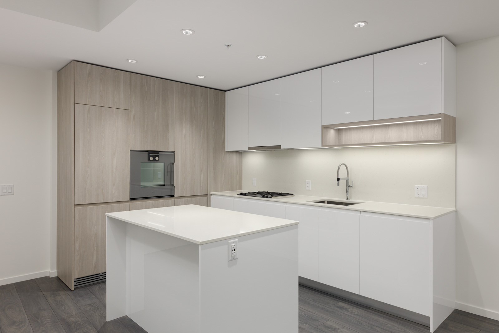Modern kitchen with white cabinets, light wood accents, built-in oven, induction cooktop, sink, under-cabinet lighting, and a white island on dark wood flooring.