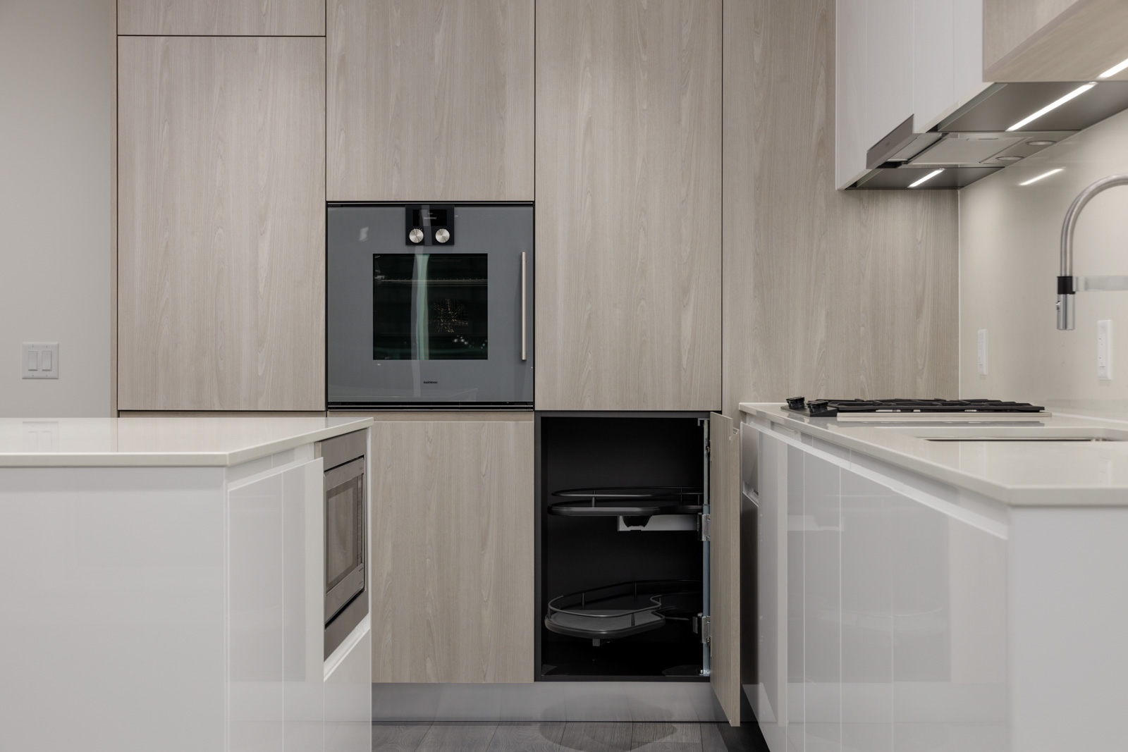 Modern kitchen with light wood cabinets, built-in oven, white countertops, induction cooktop, and an open corner cabinet showing pull-out shelves.