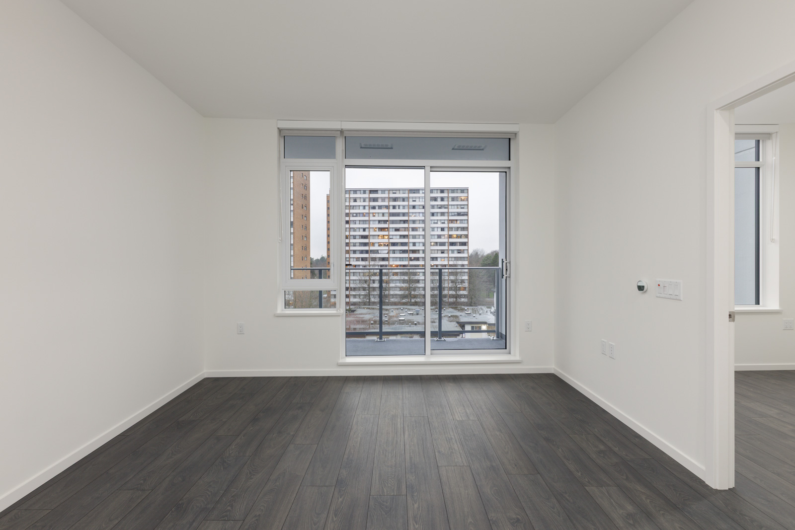 Unfurnished apartment room with dark wood flooring, white walls, a large window, and a glass door opening to a balcony overlooking a building and parking lot.