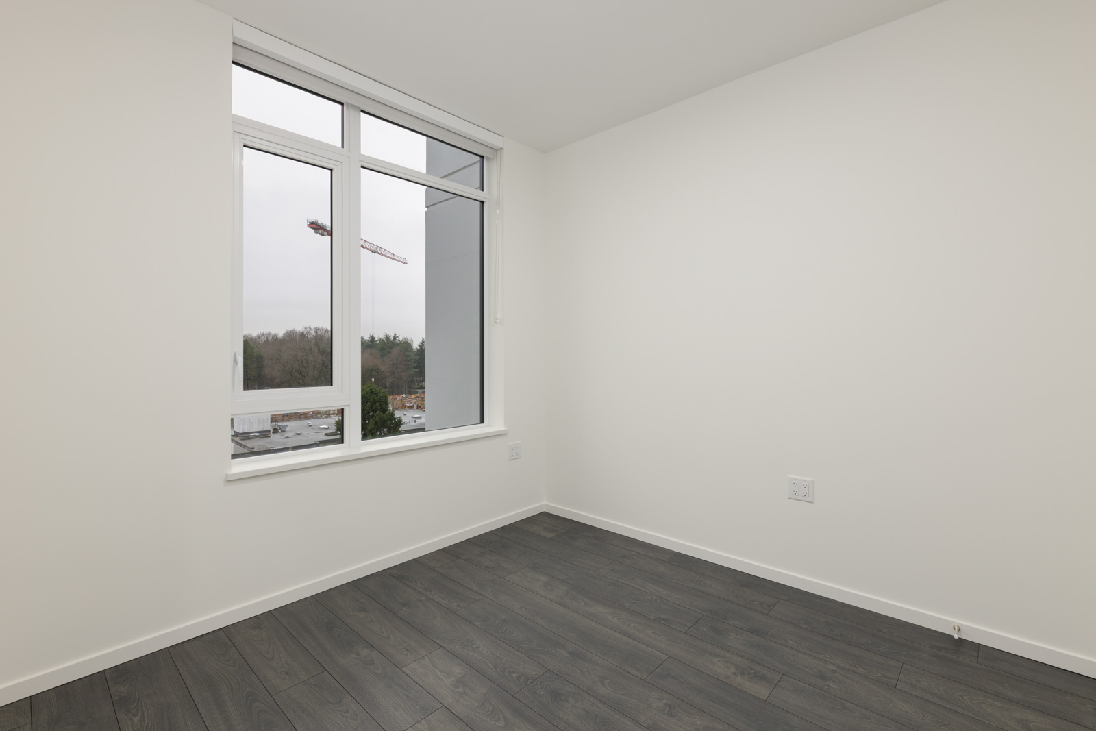 Empty room with white walls, dark wood flooring, and a large window overlooking a construction site and trees outside.