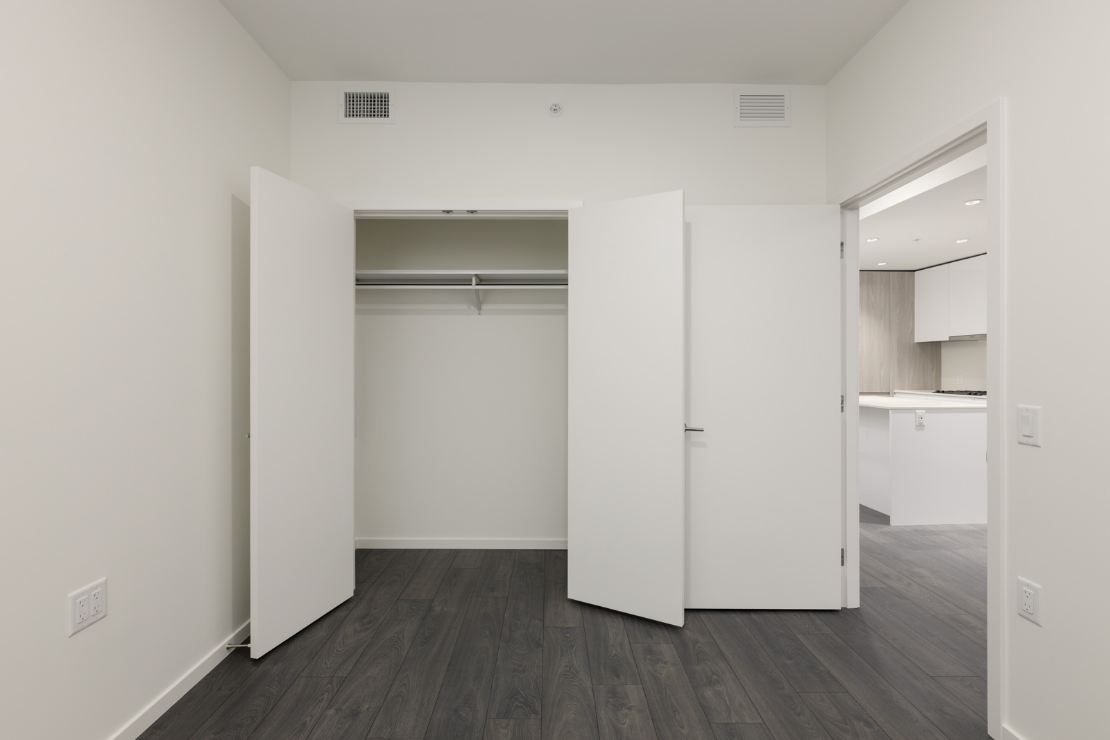 Empty room with dark wood flooring, white walls, an open closet with double doors, and an open door leading to a modern kitchen area.