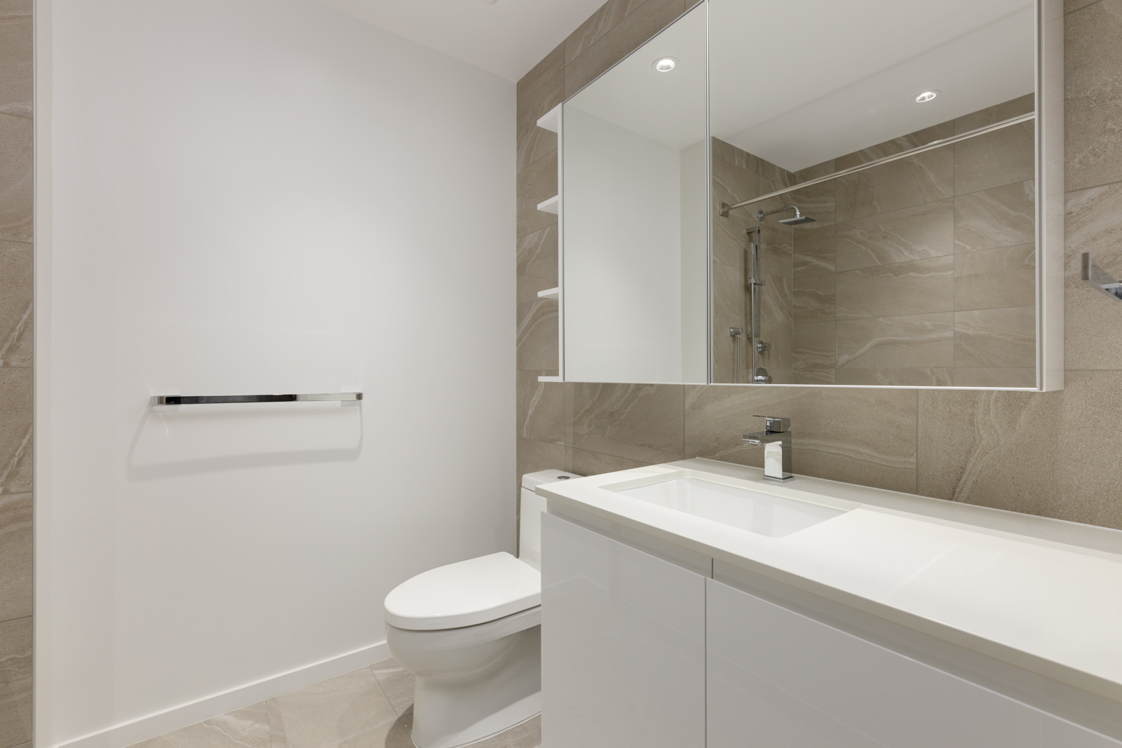 Modern bathroom with beige tiled walls, a white toilet, large mirror, white sink cabinet, and a glass-enclosed shower in the background.