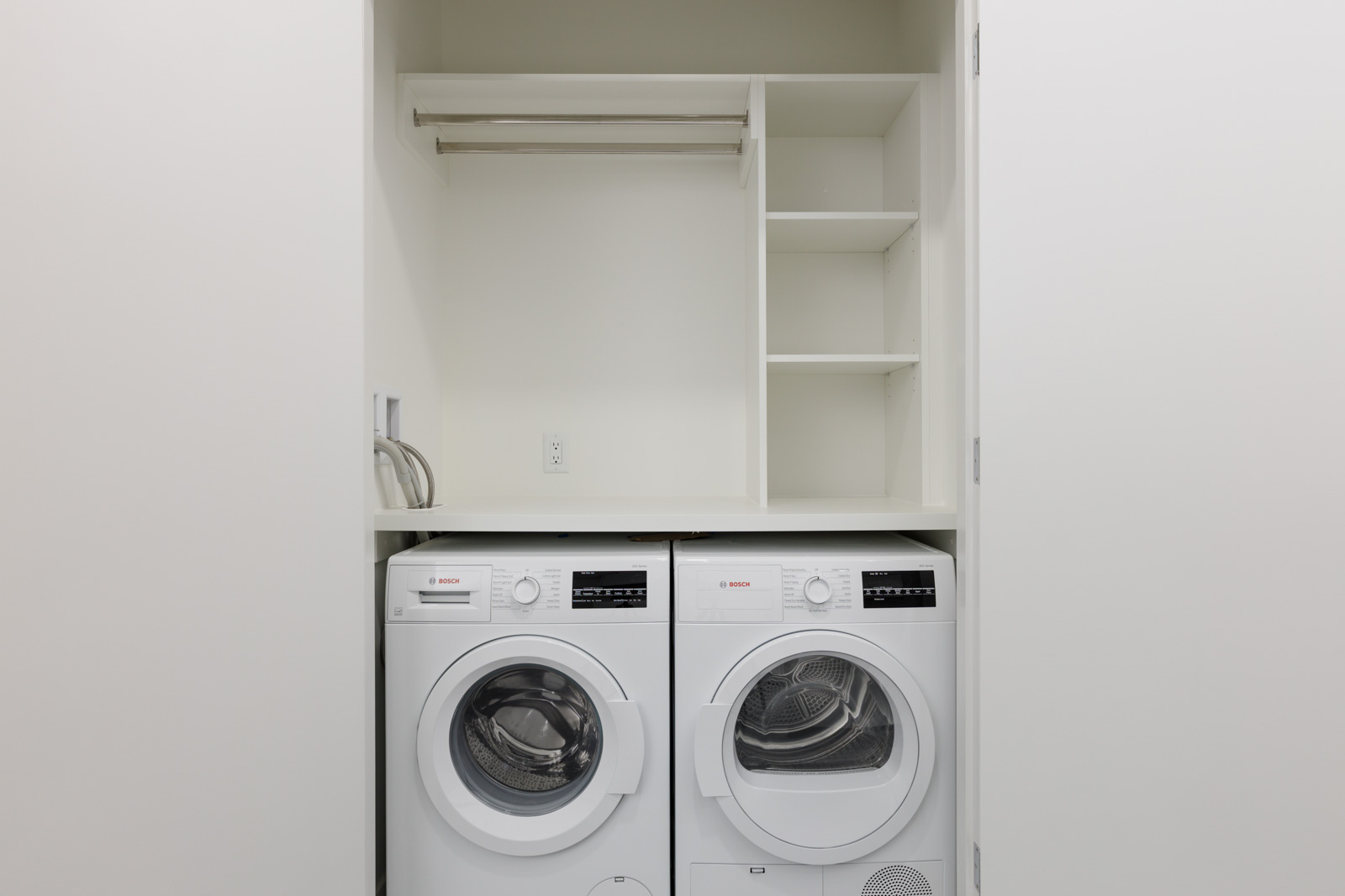 A small laundry area with a front-loading washing machine and dryer side by side, open shelving above, and a hanging rod.