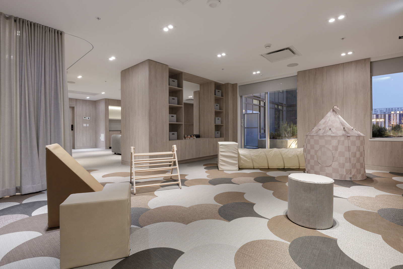 Modern playroom with neutral tones, geometric carpet, climbing structure, cushioned seating, shelves, and a small play tent near large windows.