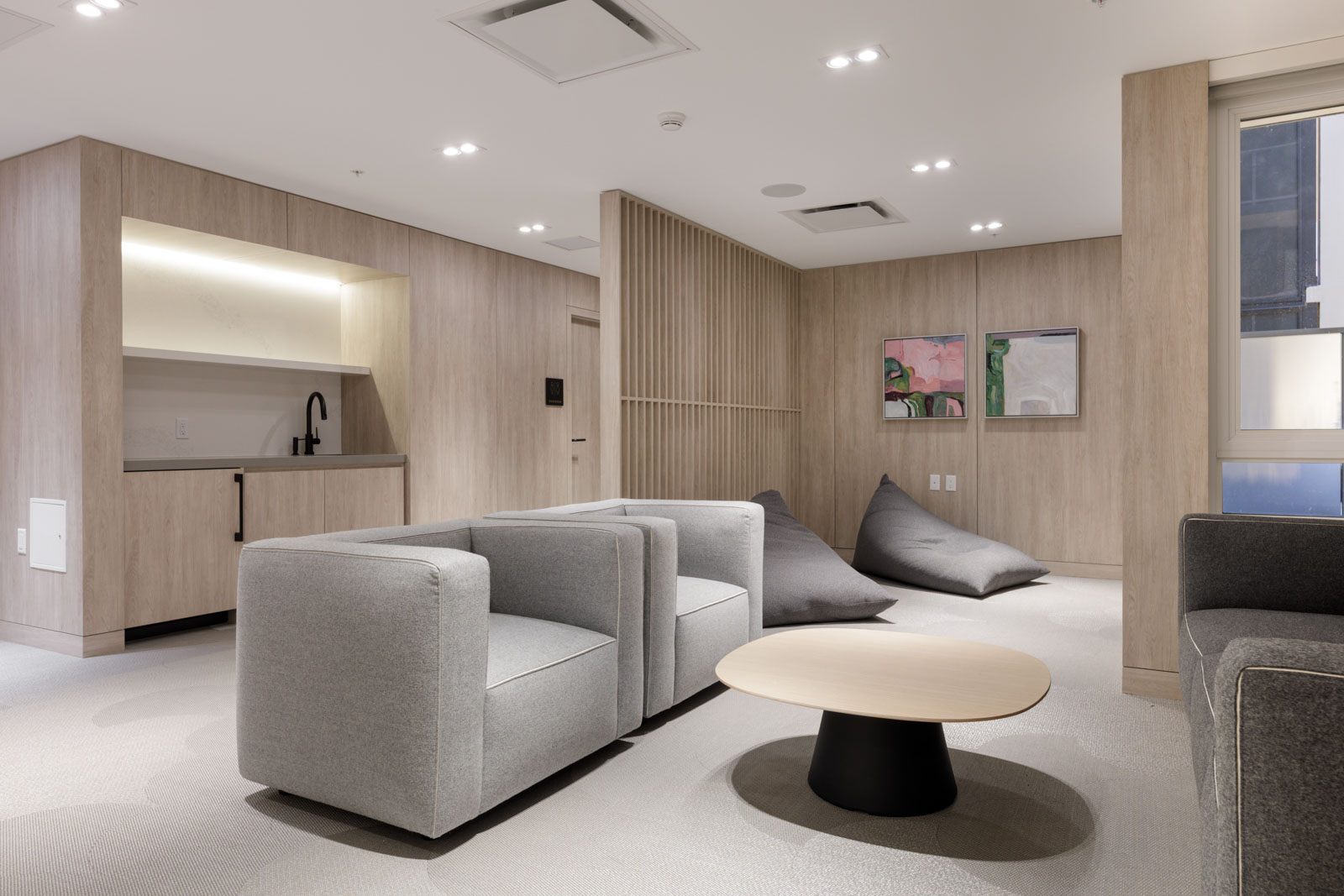 Modern lounge area with two gray armchairs, a round coffee table, large bean bags, a small kitchenette, and neutral-toned walls, featuring minimalistic decor and artwork.