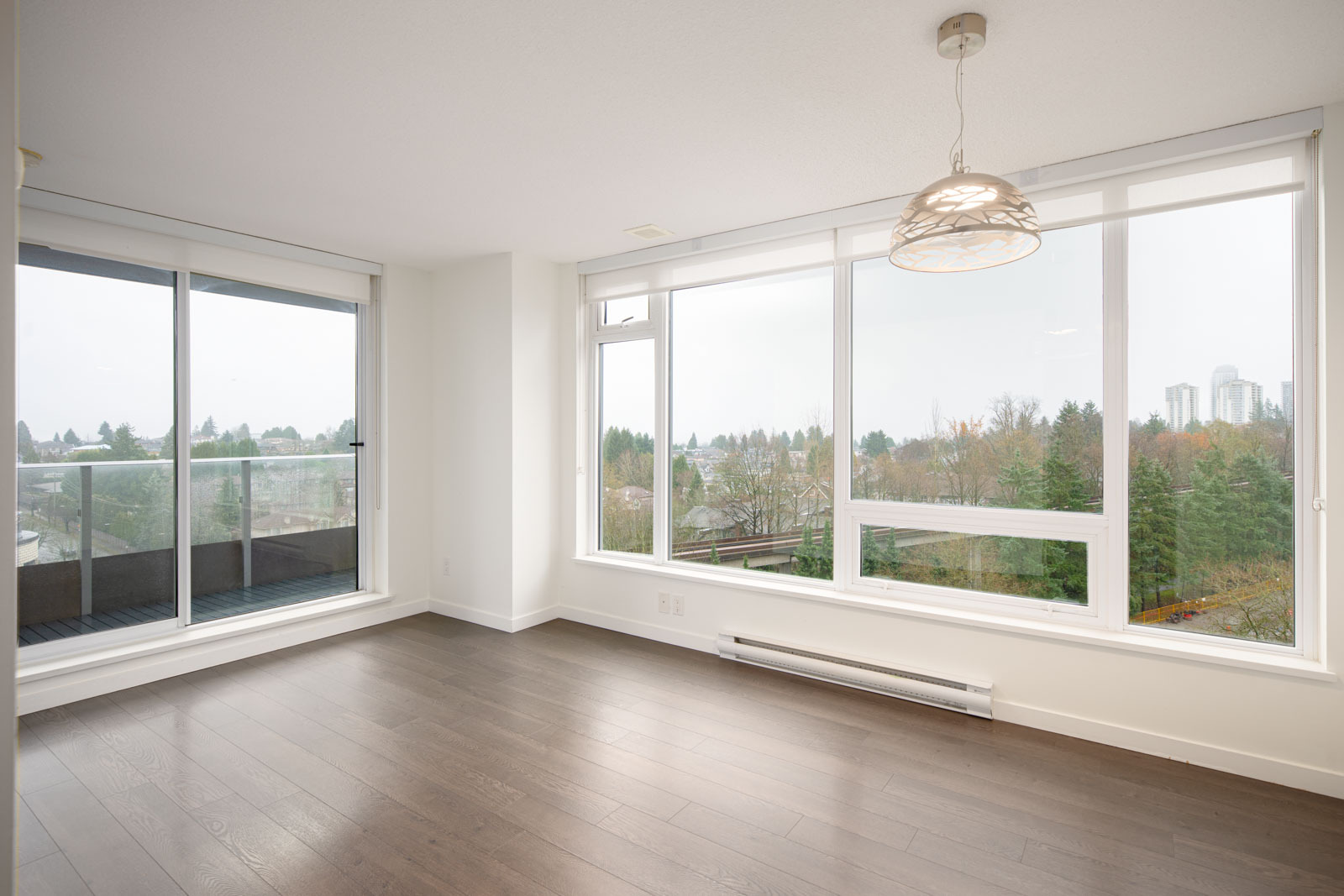 Bright, empty room with large windows and balcony access, featuring dark wood floors, white walls, and a pendant light. Trees and buildings are visible outside.