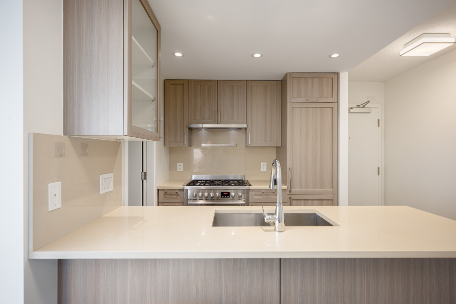 Modern kitchen with light wood cabinetry, built-in stove, stainless steel range hood, under-cabinet lighting, and a central island with a sink and faucet.