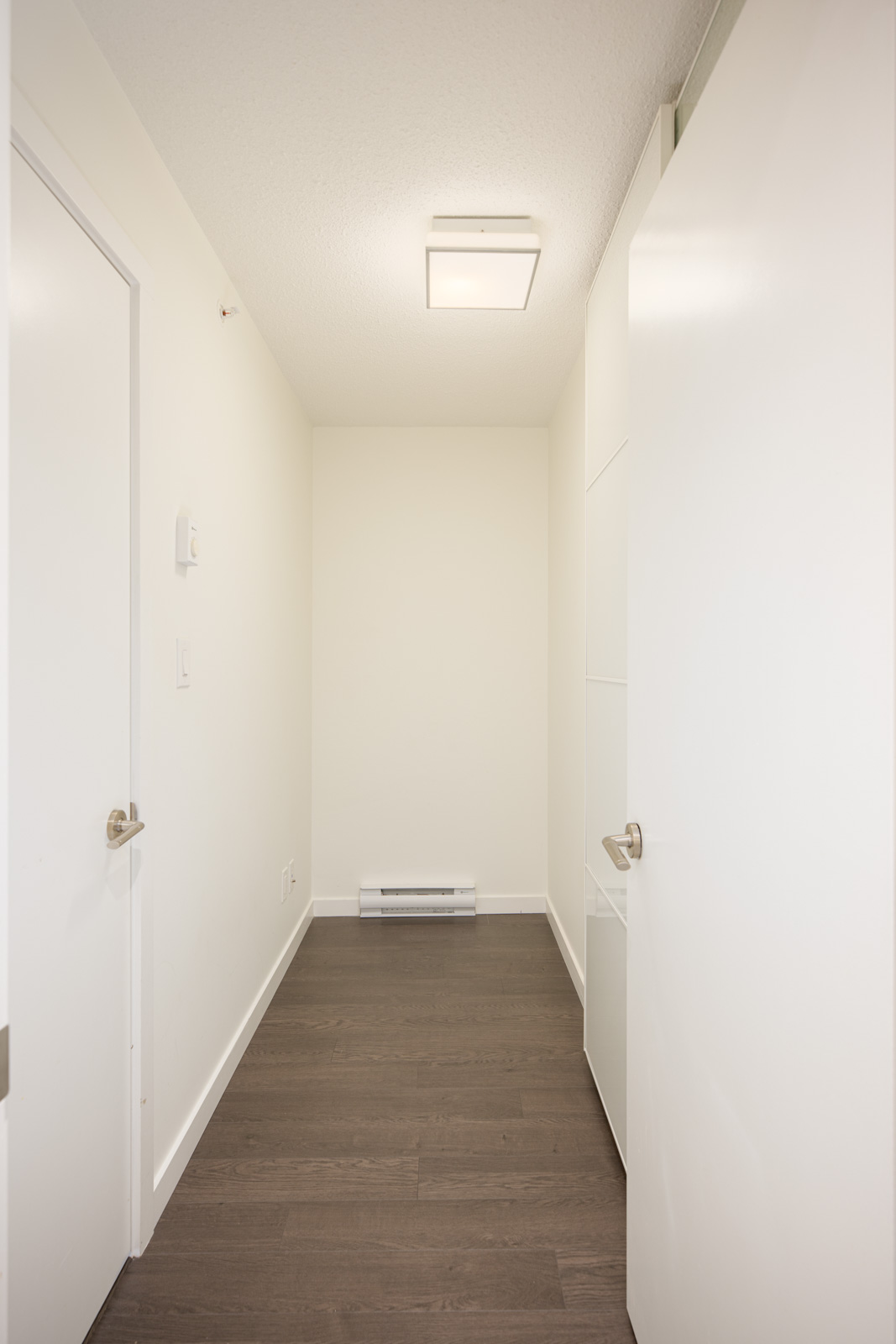 A narrow hallway with white walls, white doors, a white ceiling light, and dark wood flooring. There is a small baseboard heater at the end of the hallway.