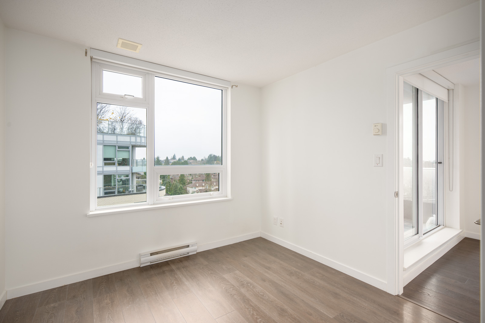 Empty room with large window, white walls, wood flooring, and a glass door leading to another area. A view of trees and a building is visible outside the window.