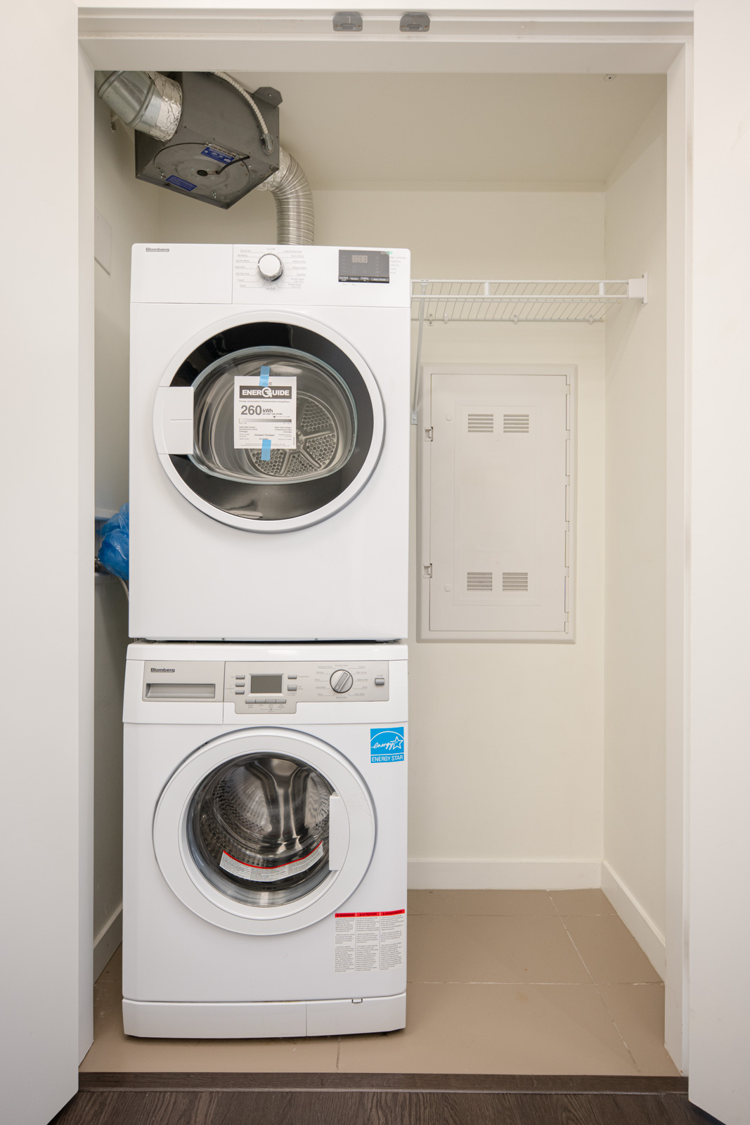 A stacked washer and dryer unit in a small laundry closet with white walls, a metal shelf, and an access panel on the right wall.