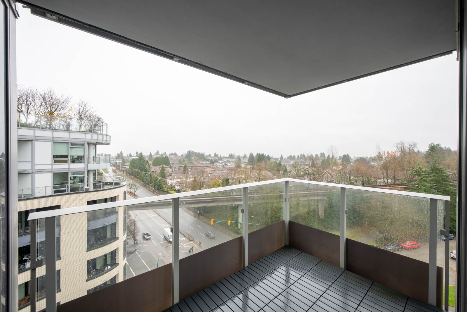 View from a covered balcony of a modern apartment, overlooking a road, nearby buildings, and trees on an overcast day.